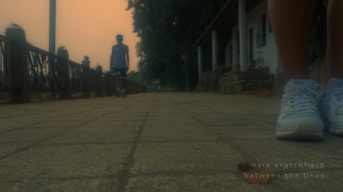 A low-angle, moody photograph of a cobblestone path at dusk, with two people walking—one in the foreground wearing white sneakers, the other silhouetted in the distance. The scene evokes movement, presence, and quiet introspection. Mark Crutchfield's “Between the Lines” is softly inscribed in the corner.