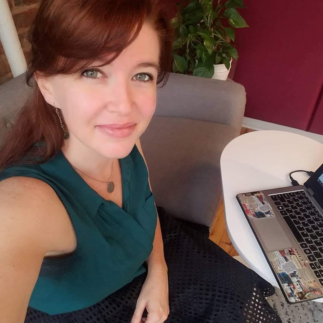 photo of a woman looking up at the camera while sitting with her laptop on a couch in a living room next to a plant. the laptop has a variety of stickers on it