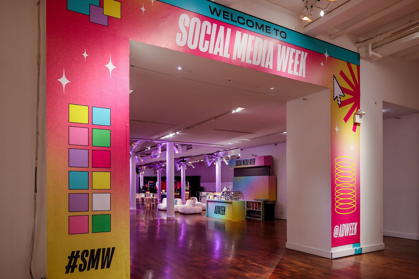 Sign welcoming people to Social Media Week SMW in New York. Background is a loft-like space with chairs and a bar branded with Adweek's name