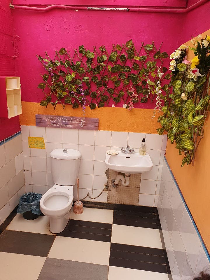 A painted wall says "#JusticiaJuanAndrés Amb L'ester", flowers are blossoming in front of it. The other image shows a small toilet bathroom with orange painted walls, flower decoration and a sign "Fascismo aquí" pointing into the toilet.
