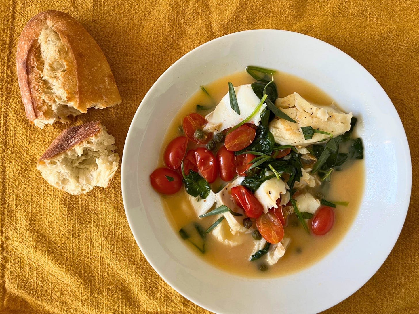 A white bowl with poached cod, spinach, tomatoes, capers, tarragon, and broth. There's some torn baguette on the side.