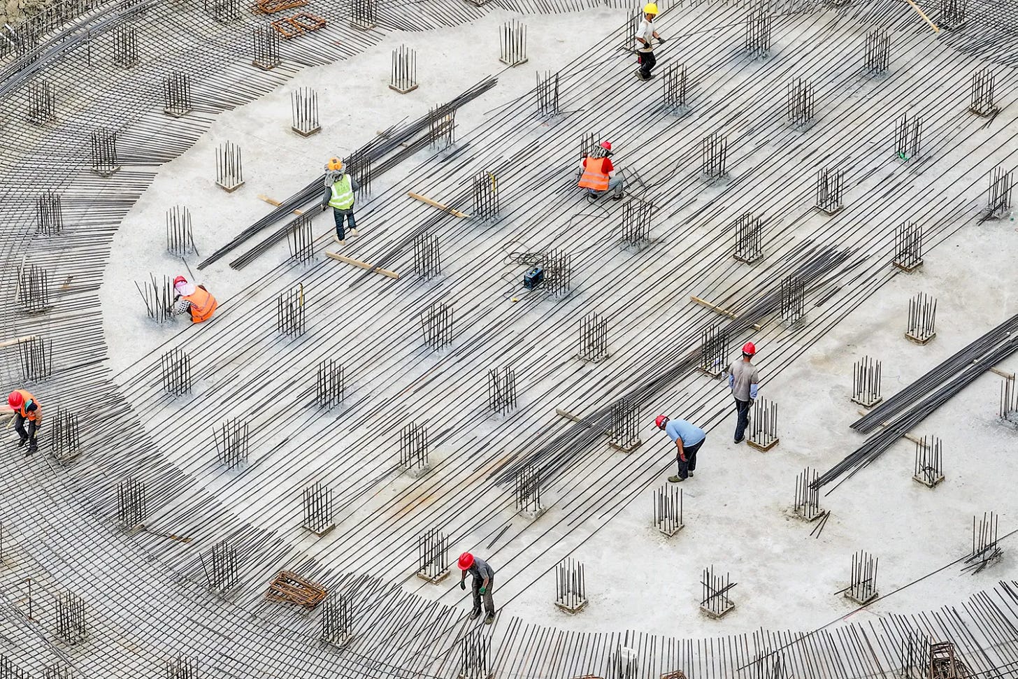 An aerial view shows several workers in hard hats and vests amid a circular rebar gridded construction site.