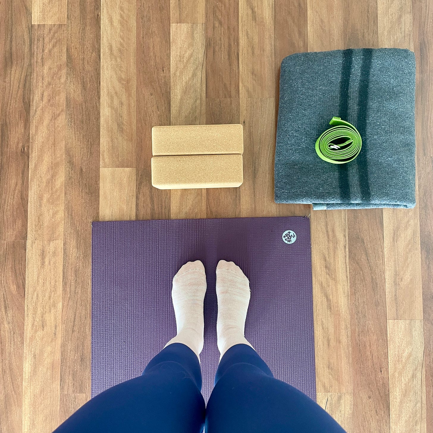 A bird's eye image of Laura's feet on her purple yoga mat with cork yoga blocks, a grey yoga blanket and green yoga strat in front of the mat. Laura is wearing blue yoga pants and light pink socks.