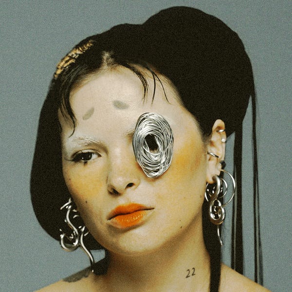 Album cover for SAYA. A headshot of Saya Gray, whose long black hair is swept into a bun, with some strands hanging down from it. She wears swirly silver earrings and a silver coil of thin metal is placed over her left eye. Her cheeks have orange blush on them and she wears coral lipstick that doesn't quite make it to the corners of her mouth. There are two, small faint eyebrows painted on her forehead. 