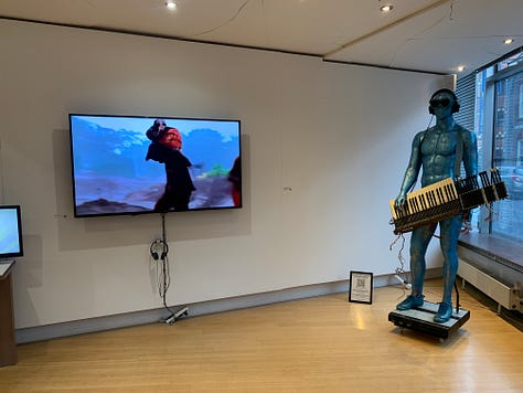 A variety of new media art works using screens, projections, including a large yellow screen and an anaologe cassete with the caption "an actual AI artist" 