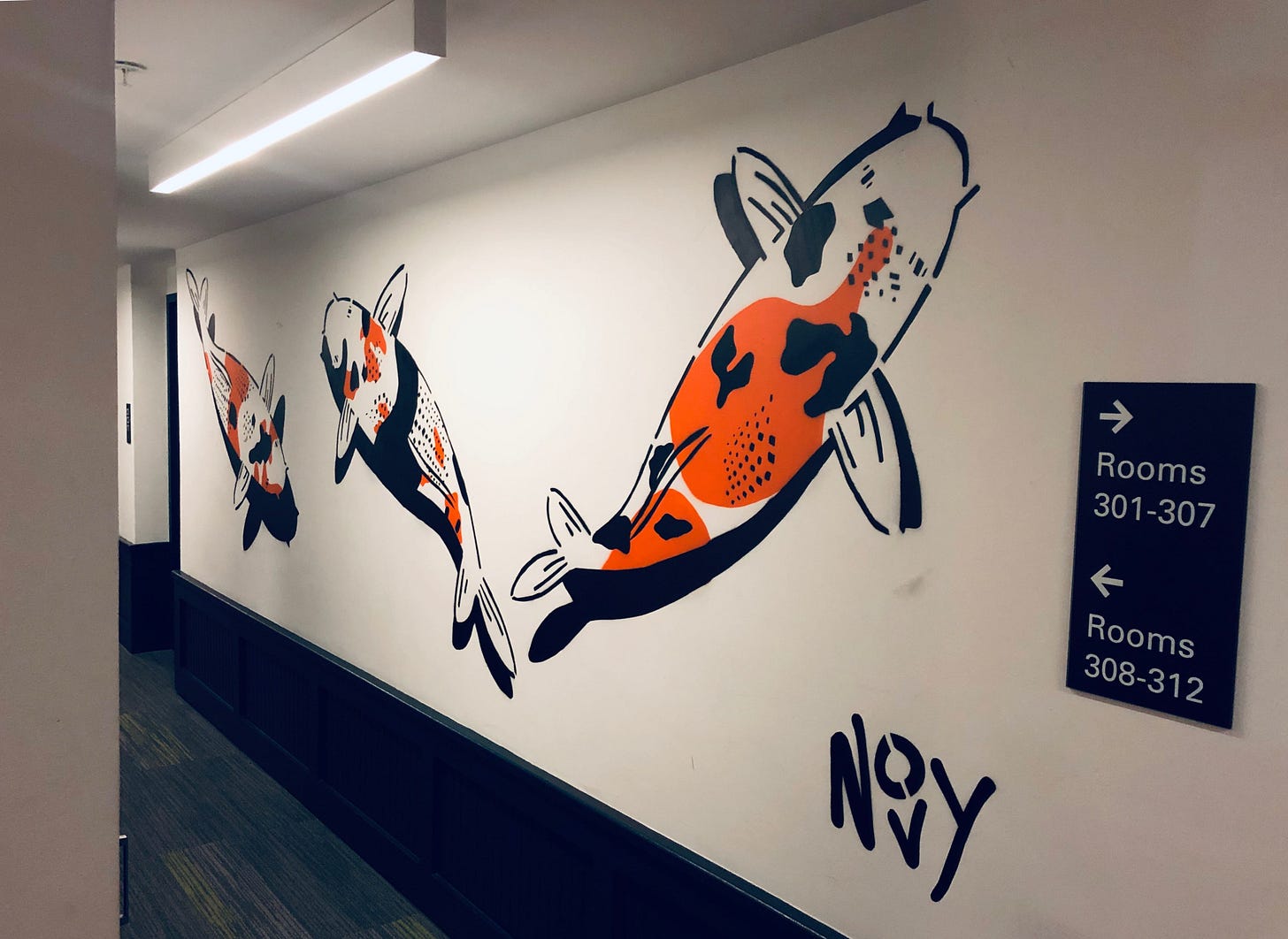 Wall mural of three stylized koi fish in black, white, and orange swimming along a white hallway with dark floors. Signs indicate directions to rooms 301-307 and 308-312. Jeremy's signature in black, Novy, is on the bottom right corner. Wall mural of three stylized koi fish in black, white, and orange swimming along a white hallway with dark floors. Signs indicate directions to rooms 301-307 and 308-312. Jeremy's signature in black, Novy, is on the bottom right corner.