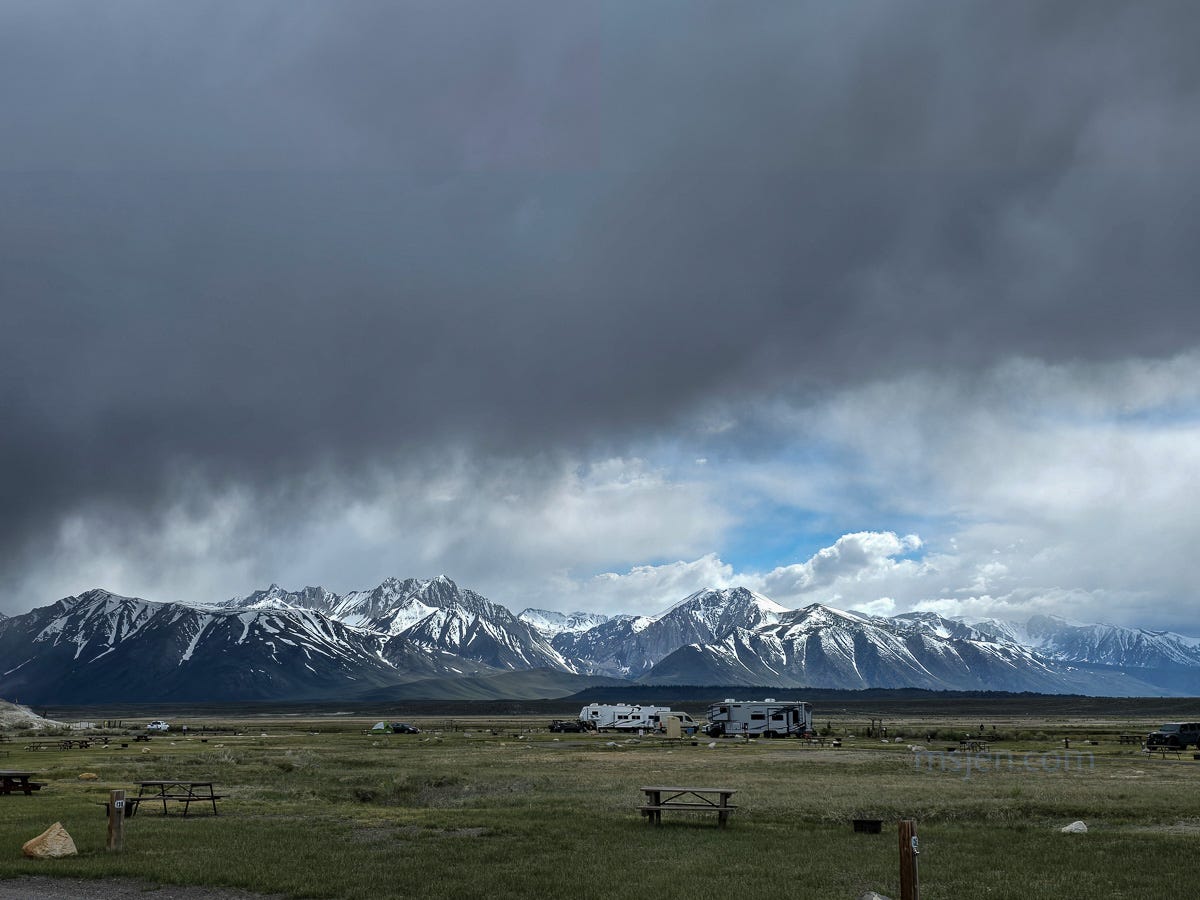 A Photo of dark gray clouds filling up the top half the image, the middle is a range of high spiky mountains with new snow on them, in the bottom of the image is a campground with a few trailers and picnic tables. Photo by Jenifer Hanen.