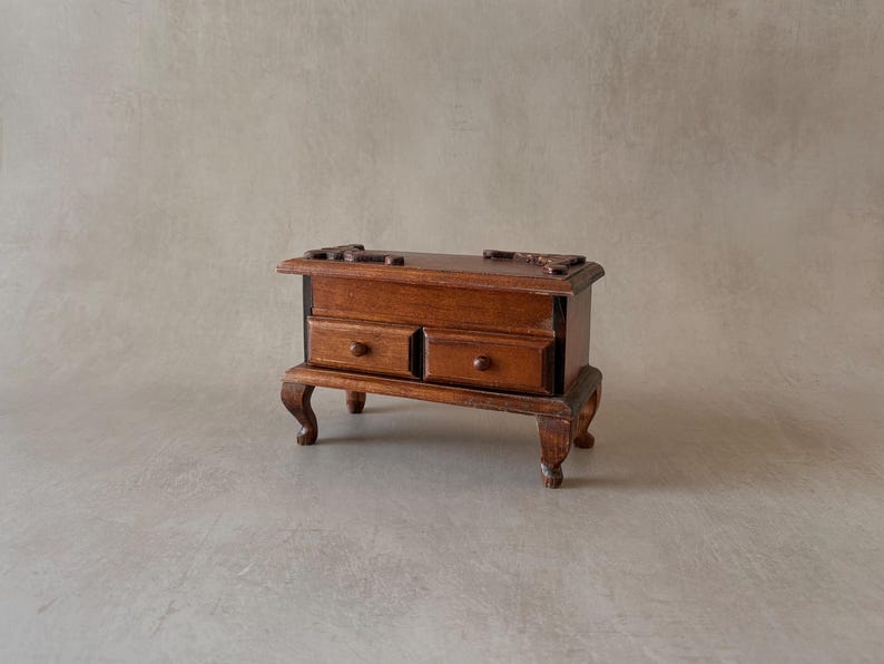 May include: A small wooden chest with two drawers and carved details. The chest is brown and has a dark stain. It sits on four legs and has a slightly distressed look.