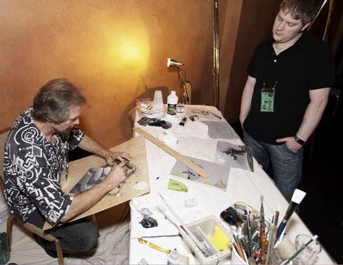 Michael Whelan works on a painting while a guest watches. The art is on a board propped at an angle between his lap and the long table that holds his supplies and other small paintings.