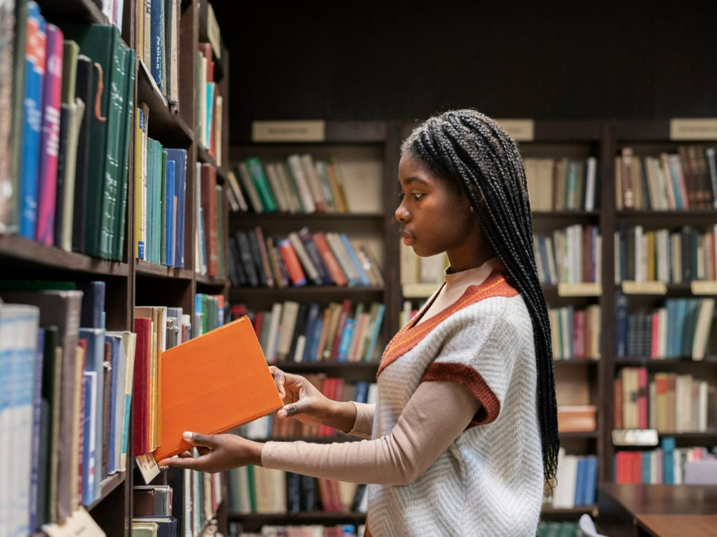 Imagem mostra uma jovem negra colocando um livro em uma estante em uma biblioteca.