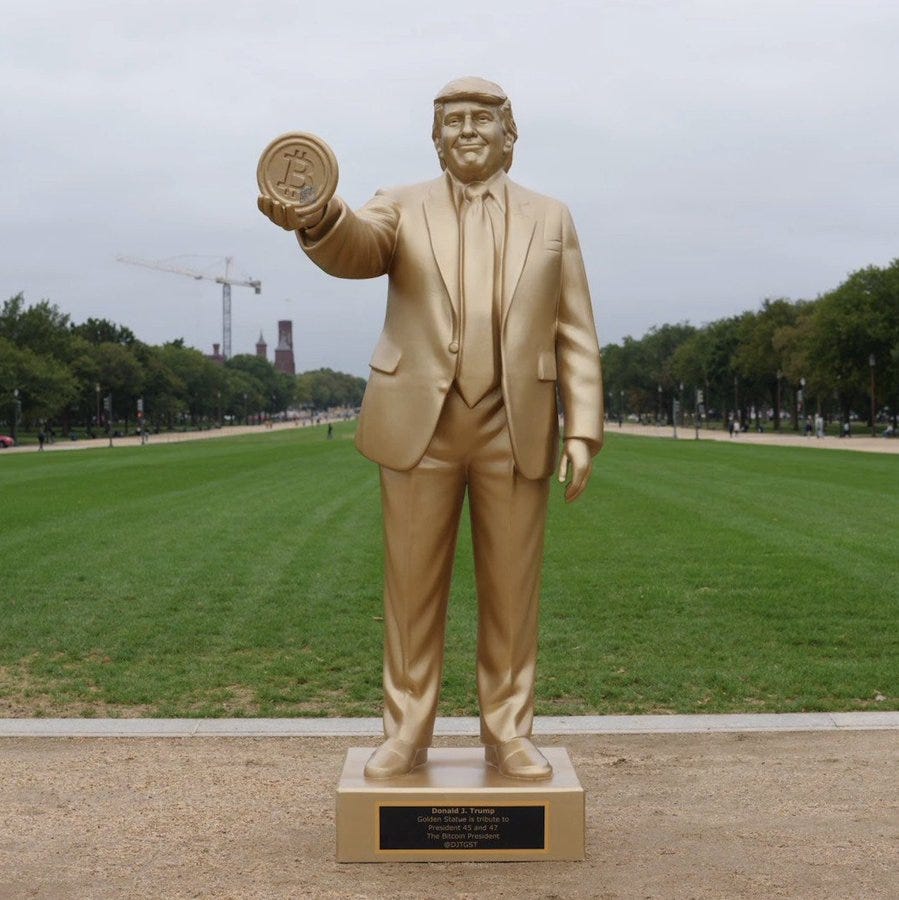 A golden statue of Donald Trump holding a Bitcoin coin. He is dressed in a suit and tie, standing on a pedestal with a plaque. The statue is positioned on a grassy field with trees and a crane visible in the background.