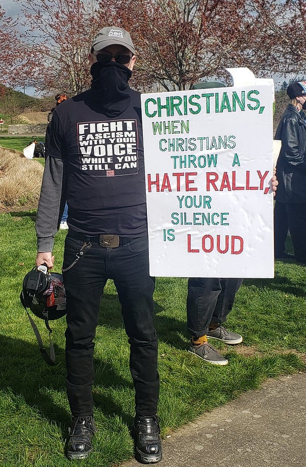 Man wearing a shirt that says, "Fight Fascism with your voice while you still can," holding a sign that says, "Christians, when Christians throw a hate rally, your silence is loud."