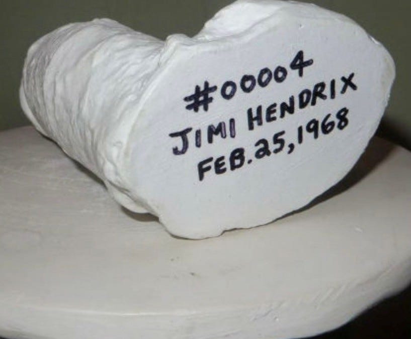 A large lump of white plaster in the shape – because that is what it was cast from – of a cock and balls. It is lying on its side on a circular while base and displaying the underside of the cast. This has written on it the words “#00004 JIMI HENDRIX FEB.25,1968”