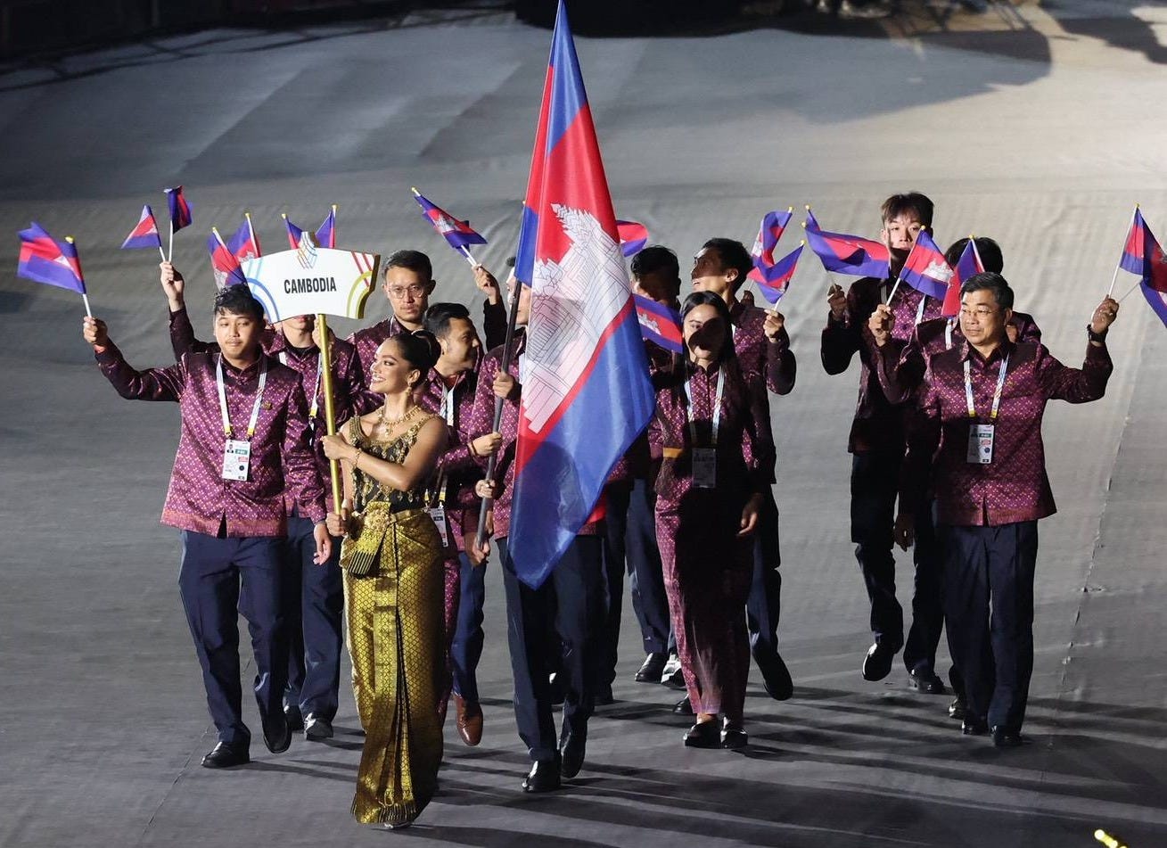 A group of athletes in regional clothing and official uniforms waved Cambodian flags and a woman carried a sign that said Cambodia. A group of athletes in regional clothing and official uniforms waved Cambodian flags and a woman carried a sign that said Cambodia.