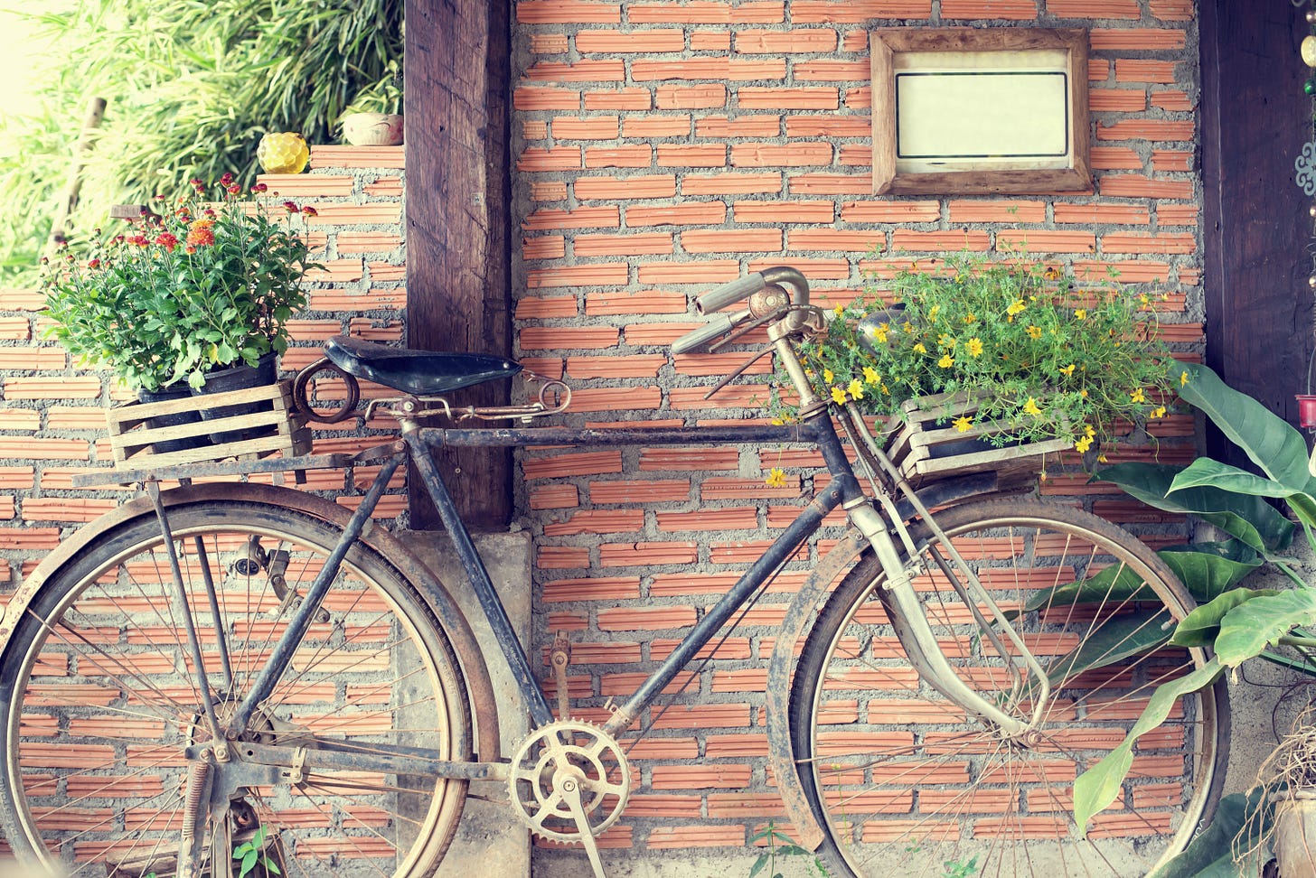 Old bicycle, rusted, resting against an exposed brick wall. It carries two wooden baskets, one at the front and another behind the seat. Both are filled with wild flowers.