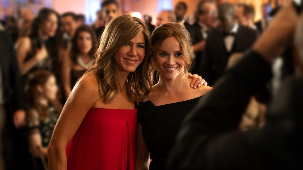 Two white women (Jeniffer Aniston and Reese Witherspoon) hug while being photographed at a fancy event