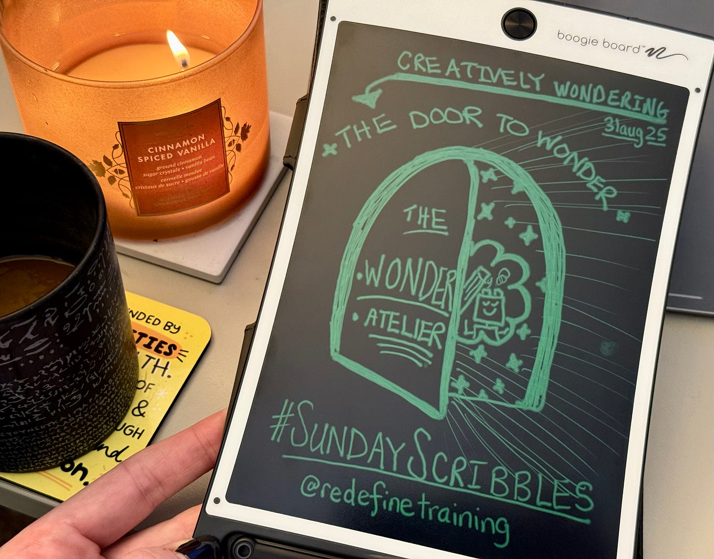 A hand holds up a Boogie Board showing a green-ink sketchnote by Georgina Dean. The drawing, titled '#SundayScribbles: The Door to Wonder,' depicts an open arched doorway labeled 'The Wonder Atelier' revealing creative icons inside. A lit 'Cinnamon Spiced Vanilla' candle and a black coffee mug are in the background.