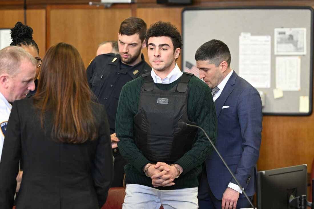 Luigi Mangione appears in court, wearing a bullet proof vest and green sweater over a while collared shirt with his hands clasped in handcuffs. Luigi Mangione appears in court, wearing a bullet proof vest and green sweater over a while collared shirt with his hands clasped in handcuffs.