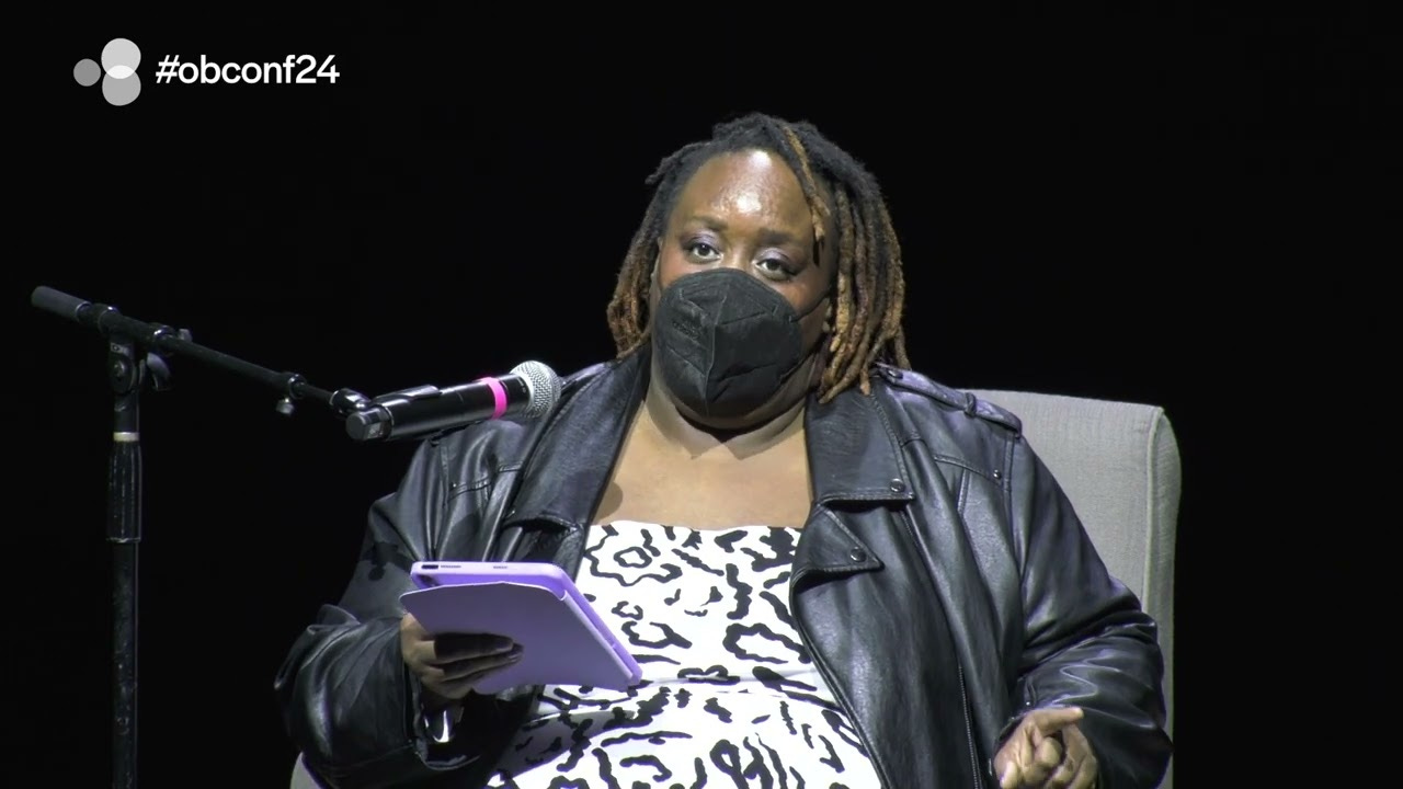 Photo de Imani Barbarin lors d'une conférence assise sur un fauteuil gris face à un micro, avc un masque ffp2 noir, un blouson en cuir noir et une robe à motifs noirs et blancs
