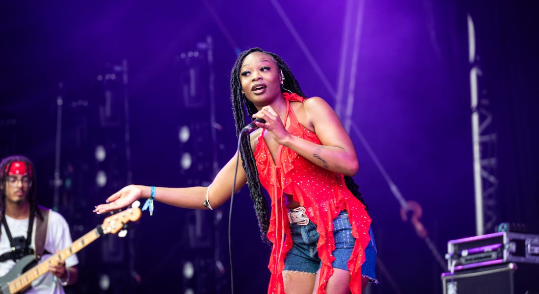 A woman wearing a red top and denim shorts, with a guitar player and purple lights behind her
