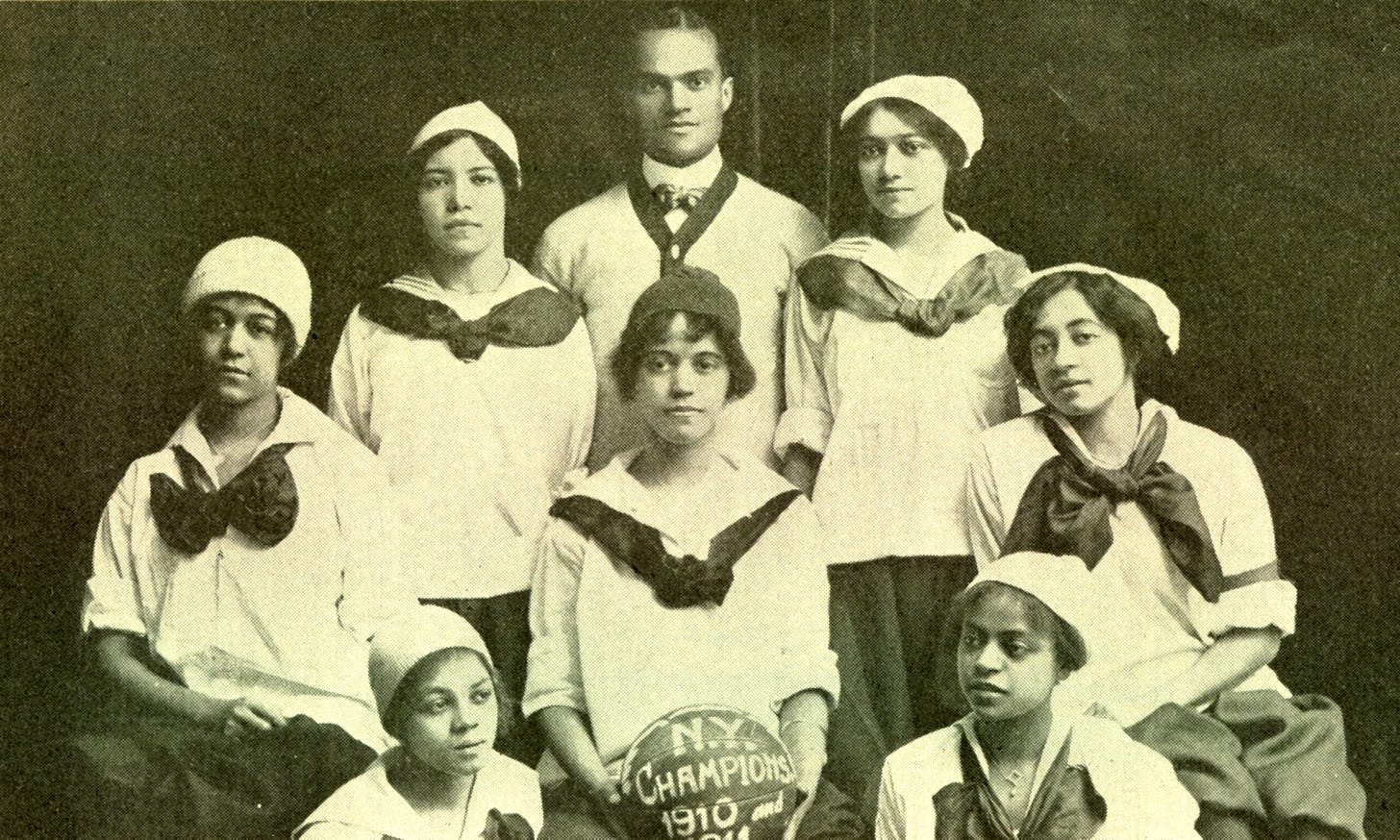 Black and white photo of seven Black women who were players on the New York Girls, all wearing white blouses with black skirts and a black bow around their necks,with one Black man who may have been their coach. One of the women is holding a basketball with the words “N.Y. Champions 1910” written on it.