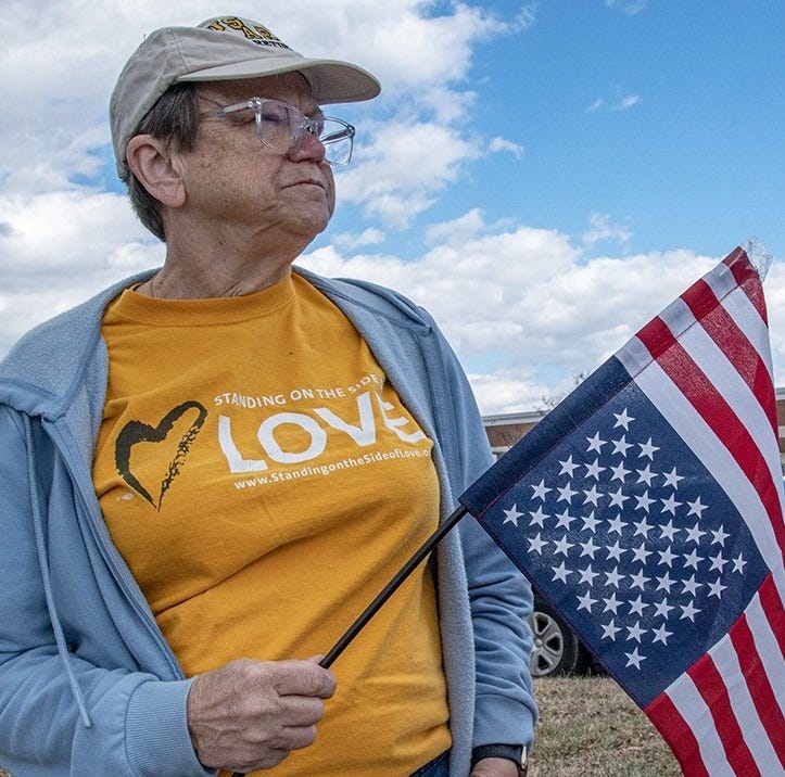 A woman wearing a "Side with Love t-shirt" and carrying a distressed (upside down) American flag