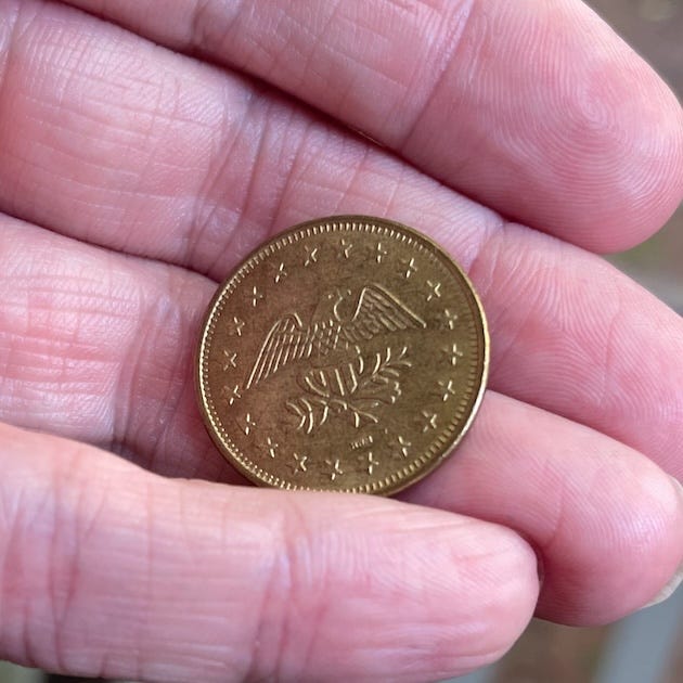 A close-up of a person’s hand holding a brass-colored token. The token features an engraved image of an eagle clutching an olive branch, surrounded by a circle of small stars. The lines and creases of the palm are clearly visible, adding warmth and texture. The background is softly blurred, keeping focus on the coin and the hand that holds it.