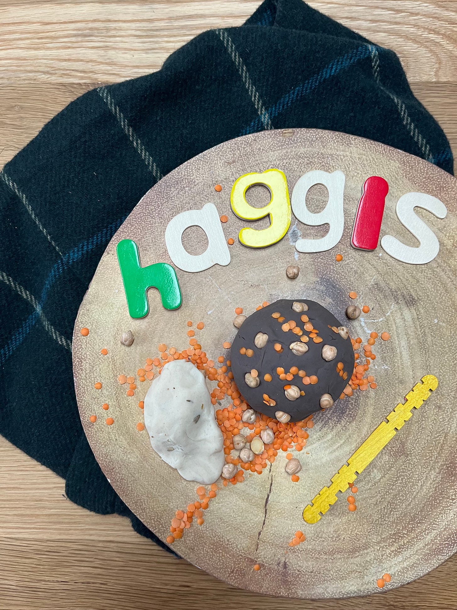 Haggis spelt out in letters, playdough and lentils for a haggis play tray. All sat on a wooden log slice with a tartan background 