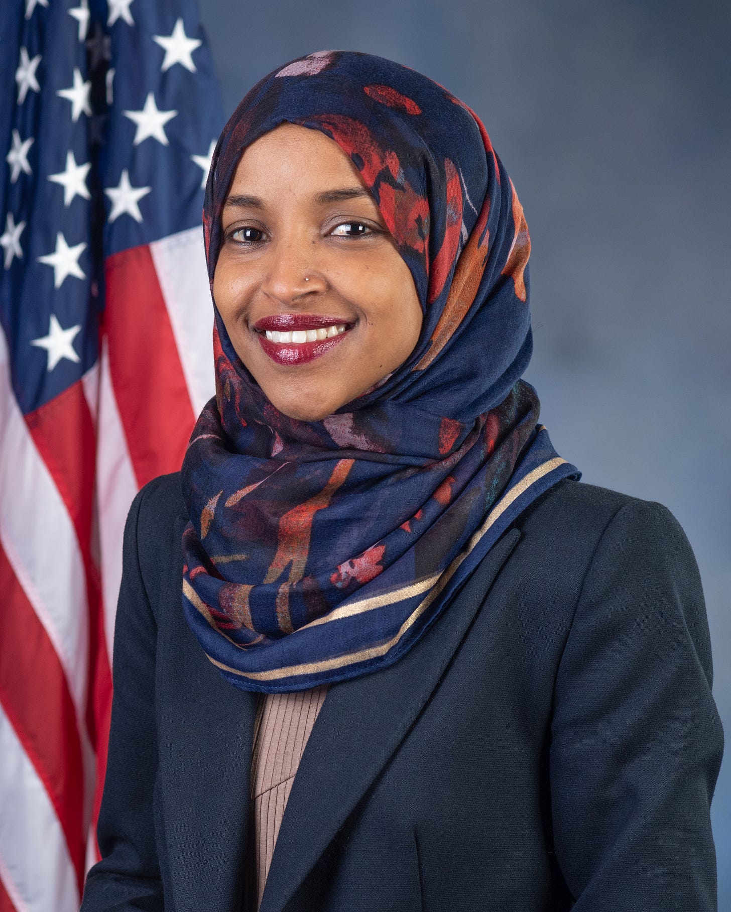 Ilhan Omar official portrait; youngish Black woman in a blue and orange headscarf and suit; behind her is an American flag Ilhan Omar official portrait; youngish Black woman in a blue and orange headscarf and suit; behind her is an American flag