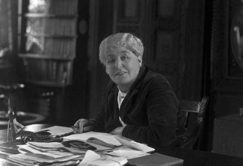 A black-and-white photo of an older white woman, with a grey short hairstyle, sitting at a table with papers in front of her. This is Maud Grieve.
