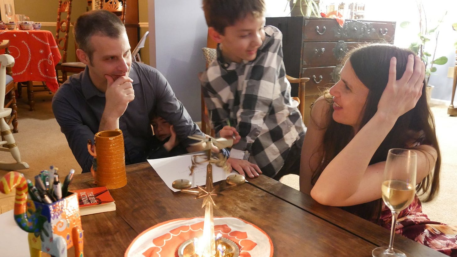 A woman and a man gaze at each other at a table. They’re smiling. There’s a boy looking at the woman with an amused expression as he is about to write something on the piece of paper. Under the man’s arm, there’s a smaller boy, looking sleepy. In front of them there’s an angel chime and some pens. Behind them there’s a dining room table with a red table cloth. A woman and a man gaze at each other at a table. They’re smiling. There’s a boy looking at the woman with an amused expression as he is about to write something on the piece of paper. Under the man’s arm, there’s a smaller boy, looking sleepy. In front of them there’s an angel chime and some pens. Behind them there’s a dining room table with a red table cloth.