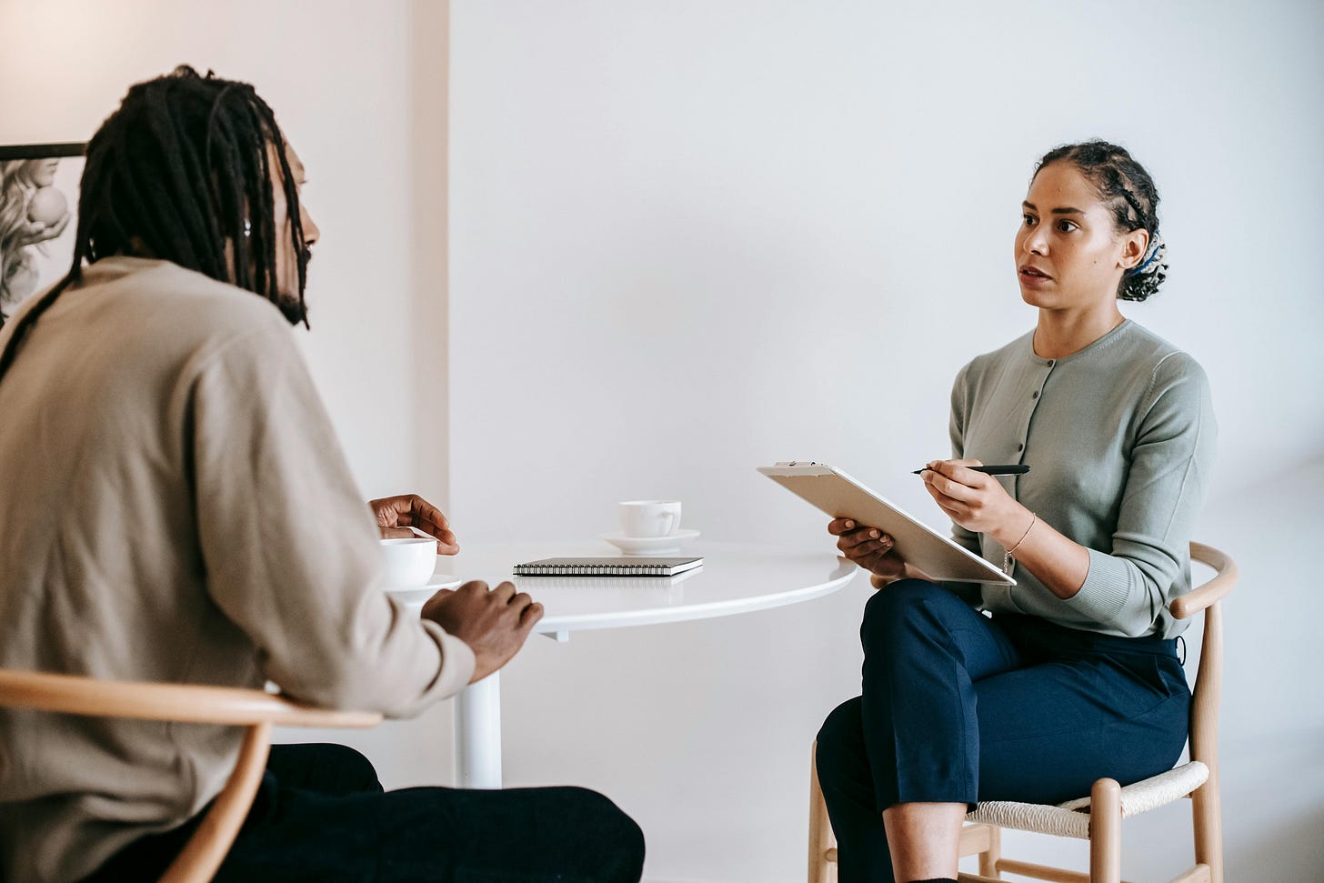 Two professionals having a one-on-one conversation at a white table, with notebooks and coffee cups, in a bright modern office setting Two professionals having a one-on-one conversation at a white table, with notebooks and coffee cups, in a bright modern office setting