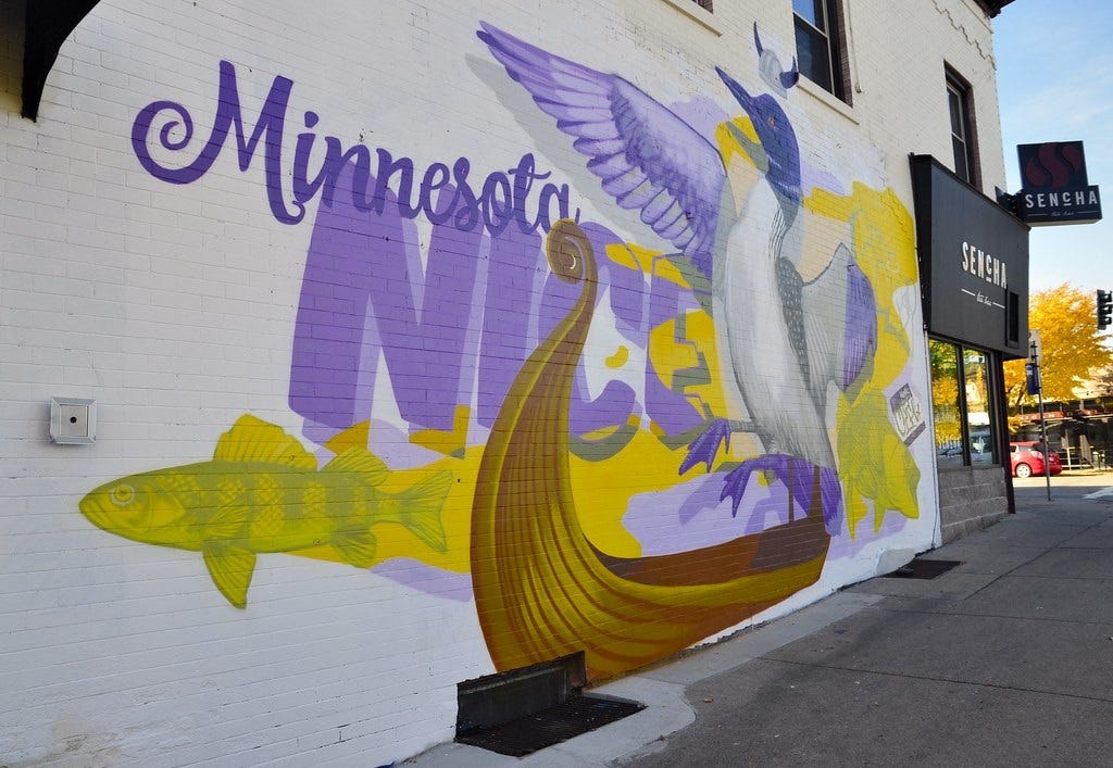 Mural on a brick building with the words 'Minnesota NICE,' and with images of a fish, a Viking longboat, a loon with its wings spread, and just above the loon's head, a Viking helmet. The mural is painted in shades of the Minnesota Vikings football team colors, purple and gold. Mural on a brick building with the words 'Minnesota NICE,' and with images of a fish, a Viking longboat, a loon with its wings spread, and just above the loon's head, a Viking helmet. The mural is painted in shades of the Minnesota Vikings football team colors, purple and gold.