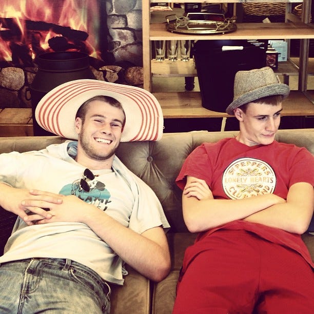 Two teen boys sit on a couch at a furniture store wearing silly hats. One grins at the camera, the other looks away in mock disdain.