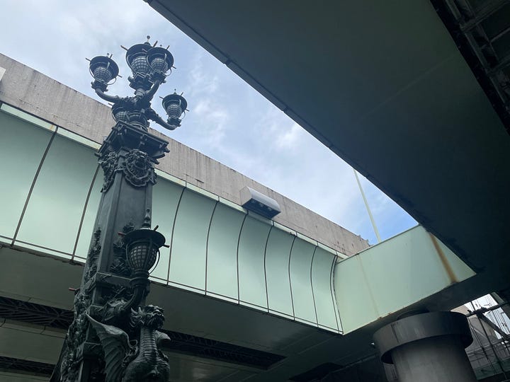 Photographs of ornate metal lanterns standing on Nihonbashi Bridge, one of which is tall and stands between the two flyover structures above, and the other is shorter and is seen with only light green metalwork above it instead of sky.