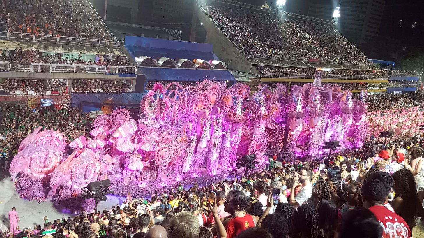 Imagem de um carro alegórico todo rosa, andando da direita para a esquerda, repleto de estátuas e pequenas rodas Imagem de um carro alegórico todo rosa, andando da direita para a esquerda, repleto de estátuas e pequenas rodas