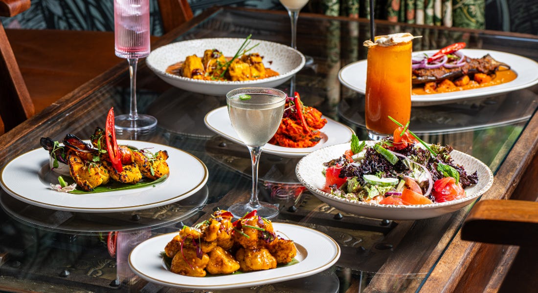 Five plates of Indian food and cocktails, on top of a glass table with wooden chairs next to it Five plates of Indian food and cocktails, on top of a glass table with wooden chairs next to it