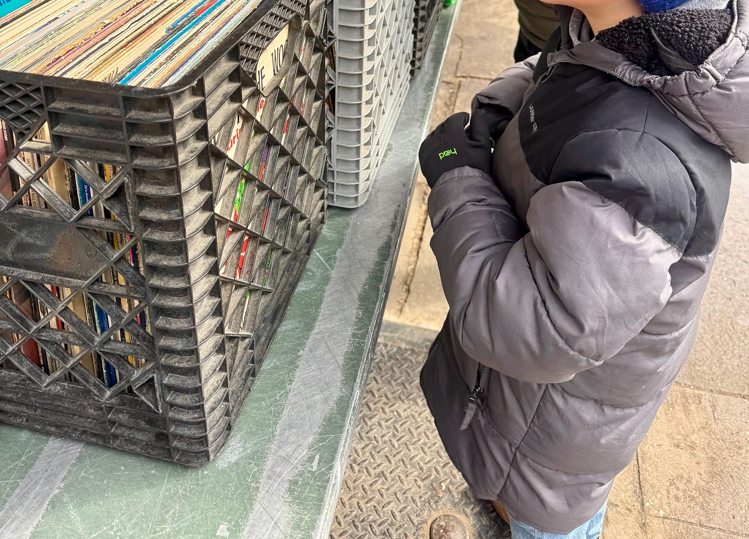 A little boy in a grey puffer coat and wearing black gloves stands in front of milk cartons filled with records on a set on a table on a sidewalk