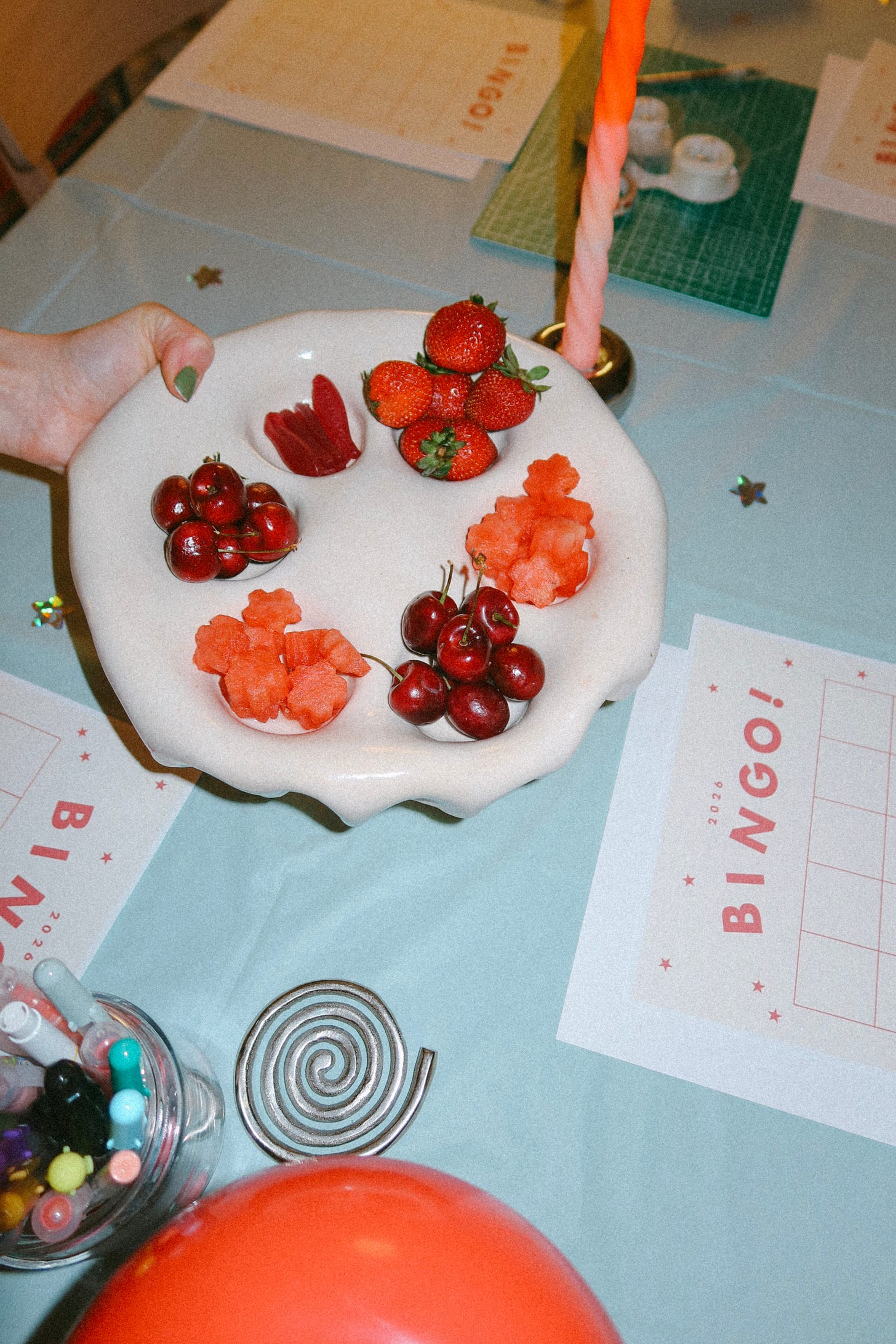 Hand holding a scalloped plate of strawberries, cherries, and flower-shaped watermelon on a table