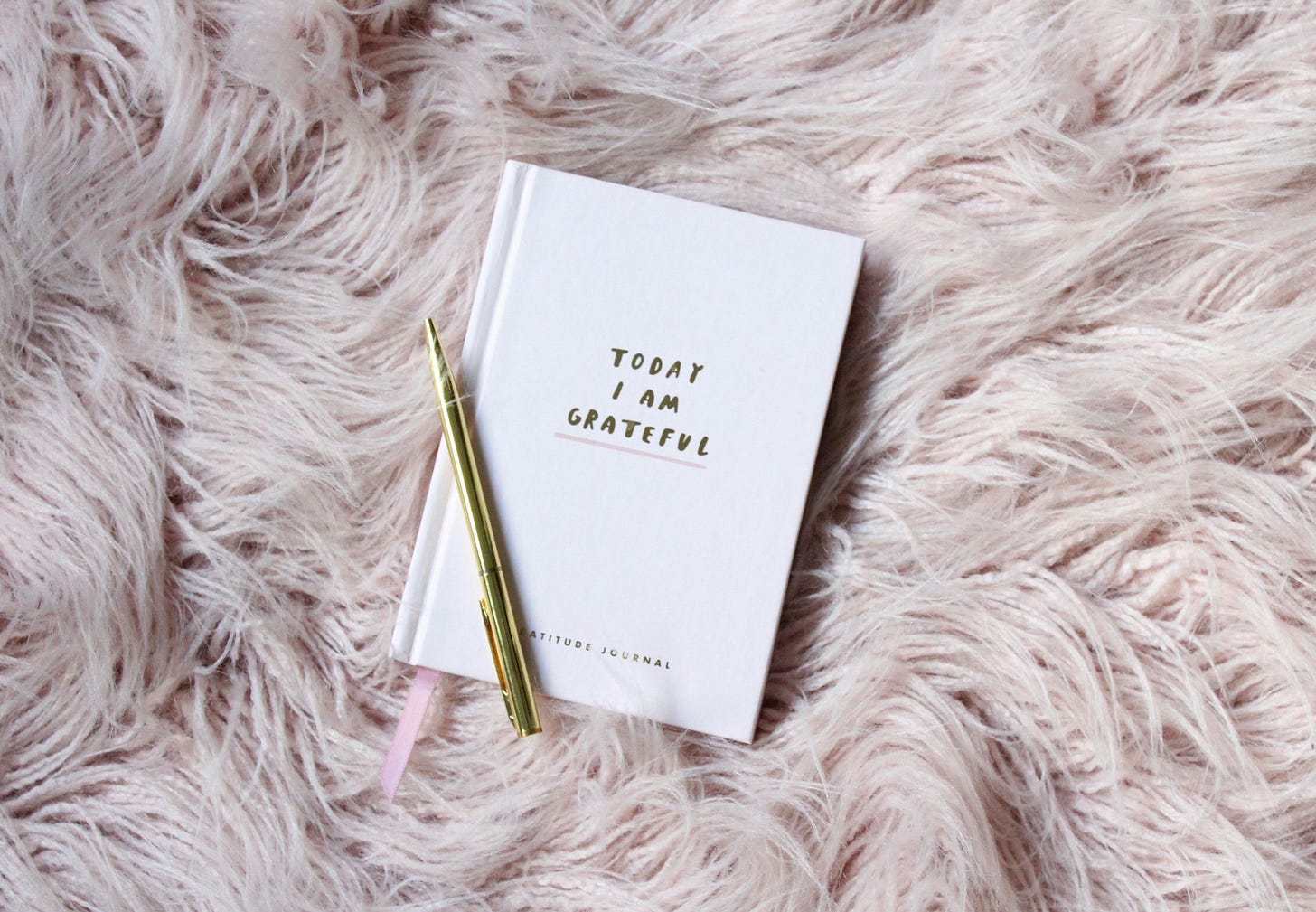 A white gratitude journal with gold lettering and a gold pen rests on a soft pink faux-fur surface. Photo by Gabrielle Henderson on Unsplash