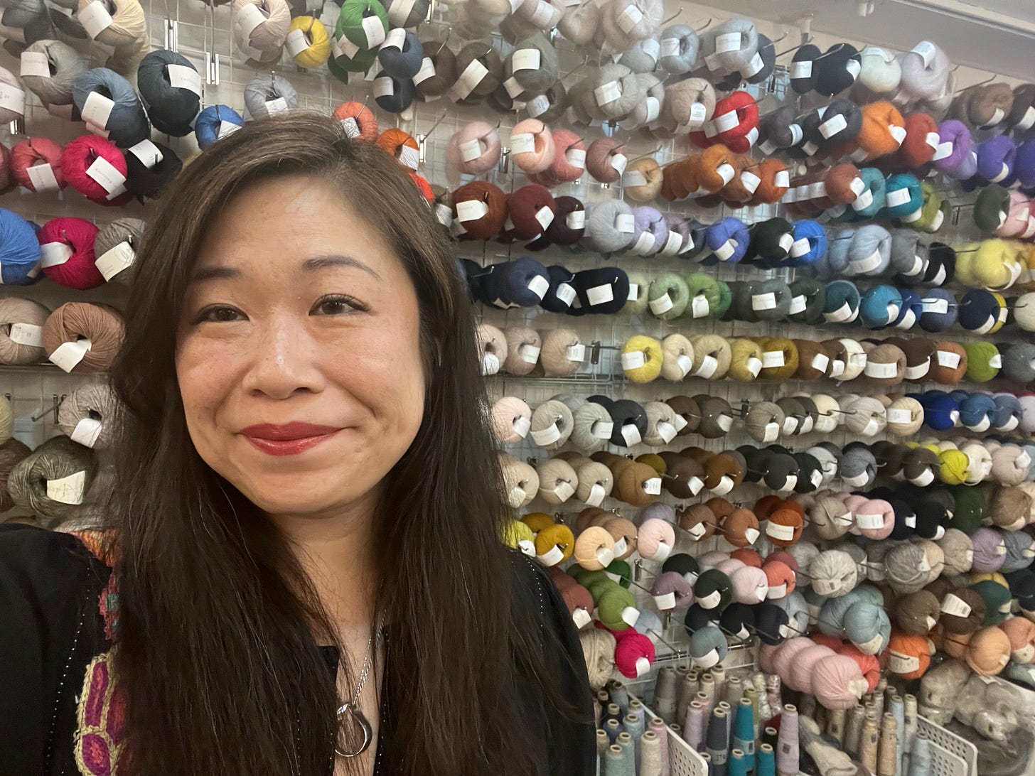 East Asian woman dressed in black, with long hair and red lipstick, poses in front of a wall filled with balls of yarn on racks East Asian woman dressed in black, with long hair and red lipstick, poses in front of a wall filled with balls of yarn on racks