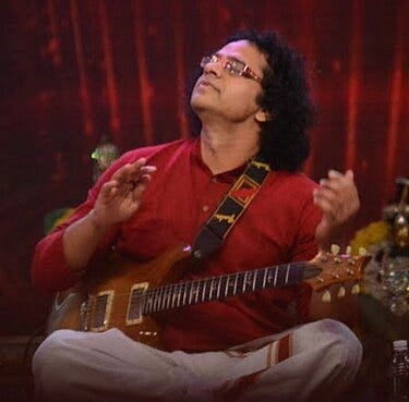 Picture of Prasanna, , wearing a red kurta and light-colored pants, seated cross-legged on stage while playing an electric guitar. He looks upward with eyes closed, appearing deeply immersed in the music. Picture Credit: Wikipedia