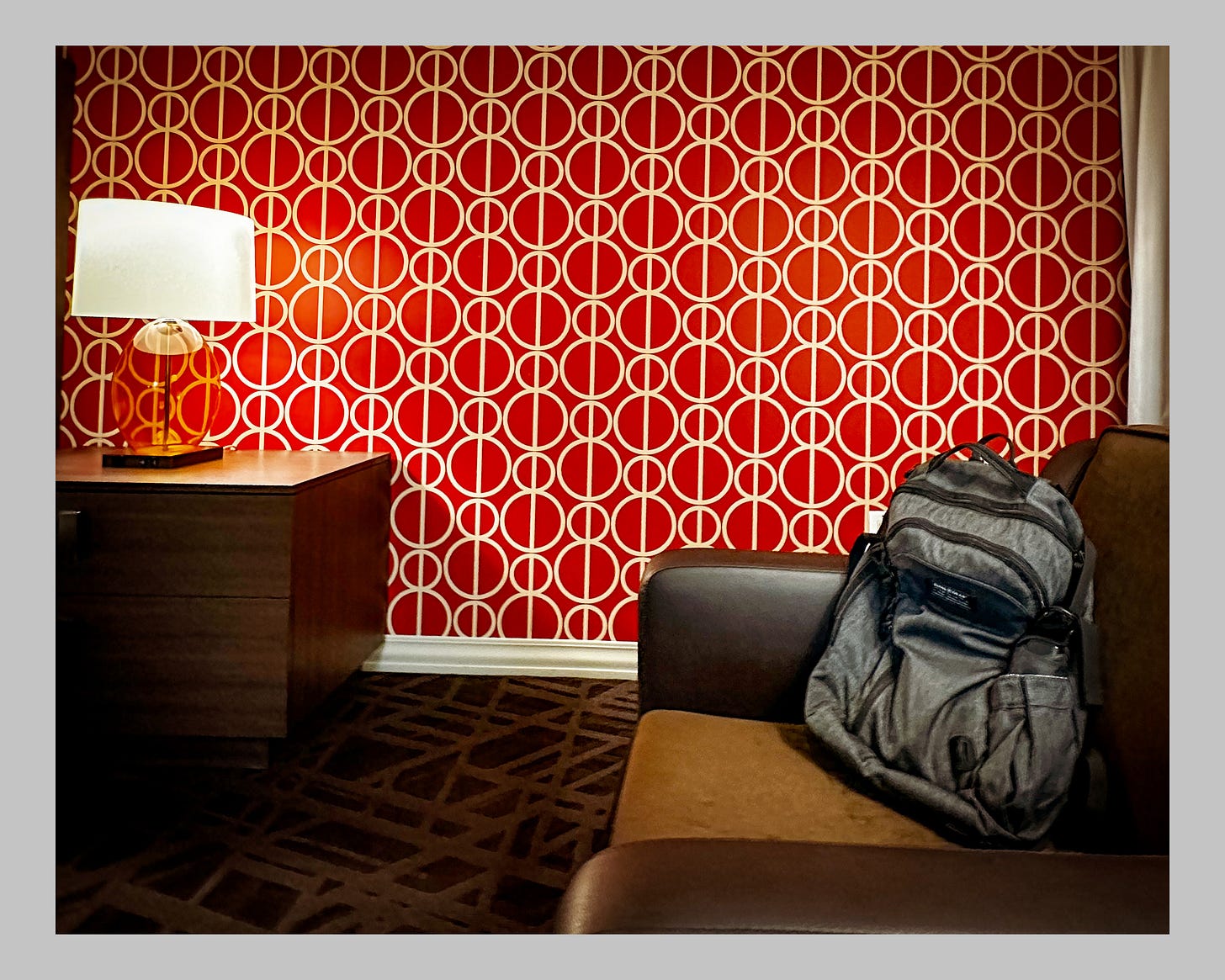 A hotel room with mod-style wallpaper. A gray backpack sits in a brown leather chair.