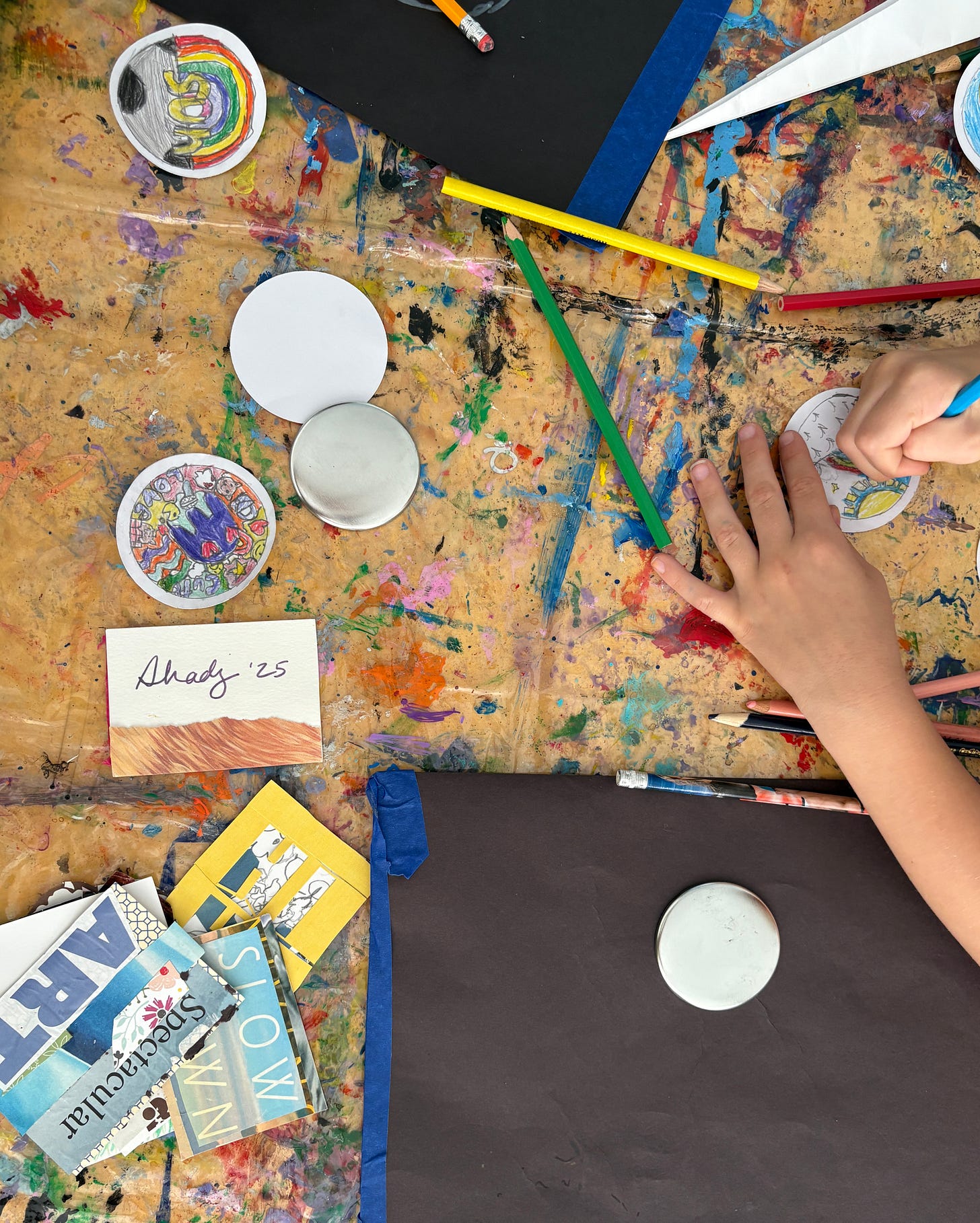 A students hands drawing a button. On the table are several other colorful buttons and collaged artist trading cards.