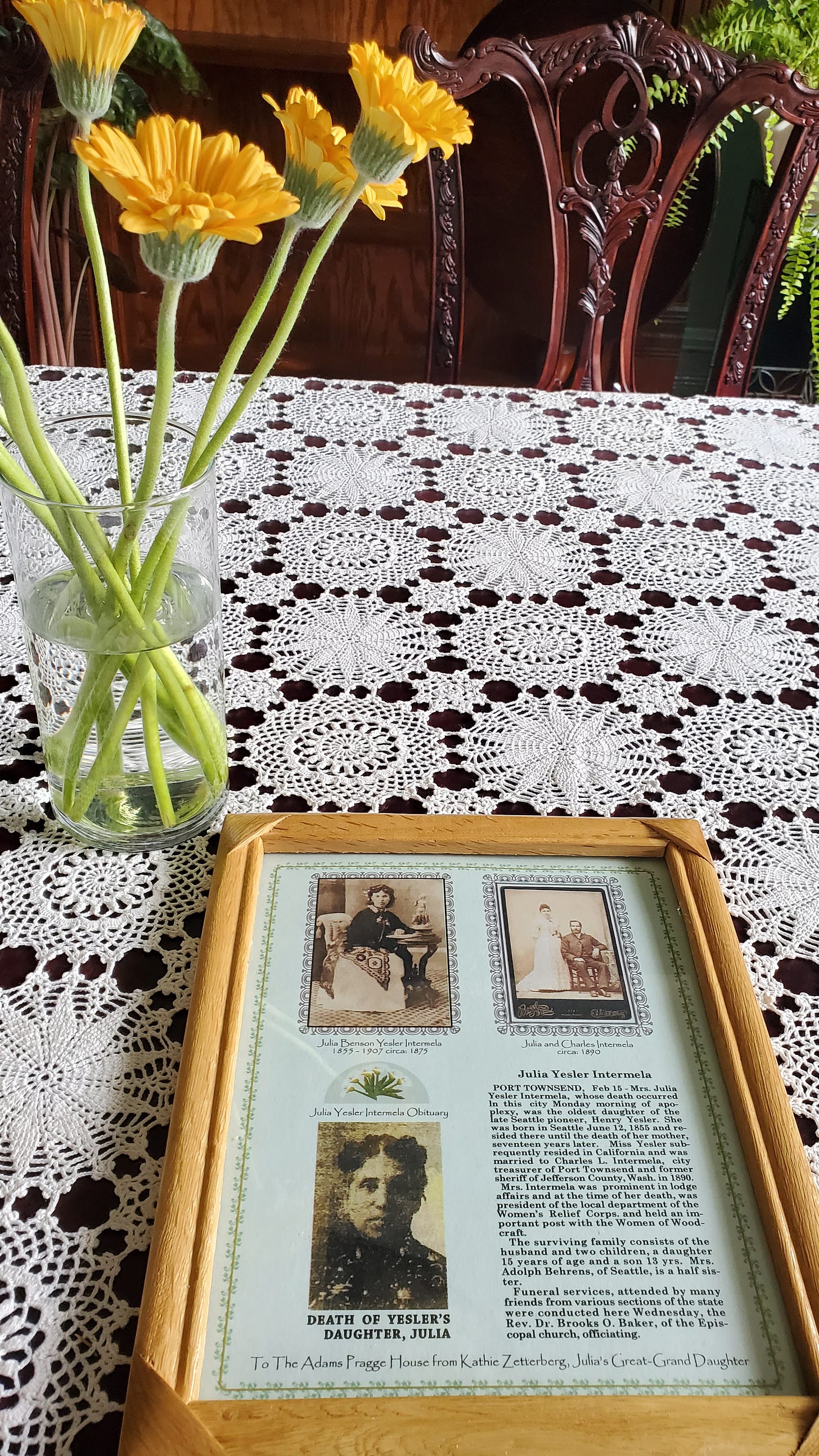 Framed display containing photographs and obituary clippings of Julia Yesler Benson Intermela, placed on a lace-covered table.