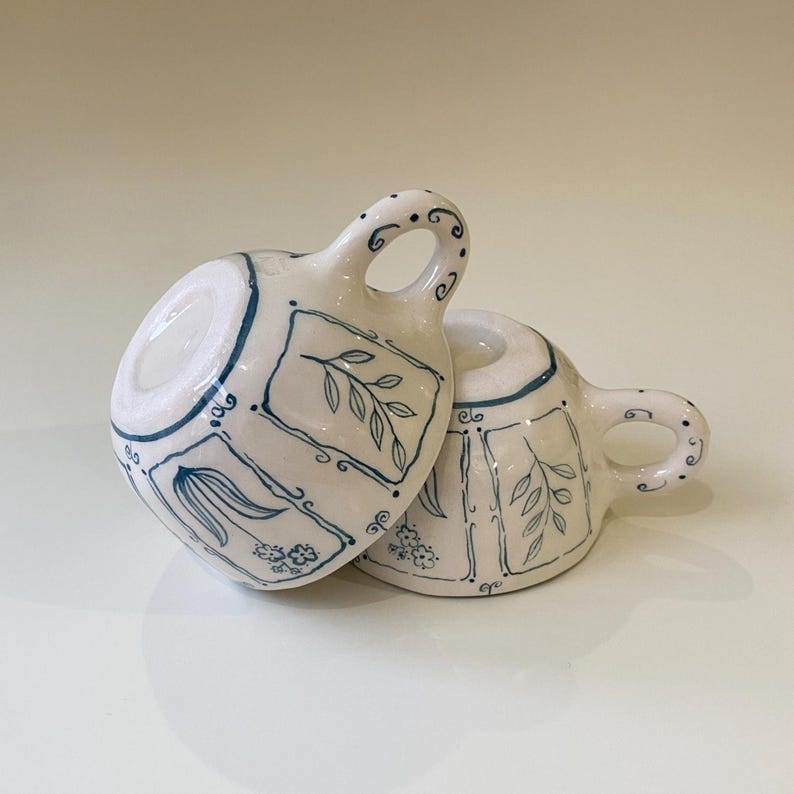 May include: Two white ceramic teacups with blue floral designs. The cups are stacked on top of each other.