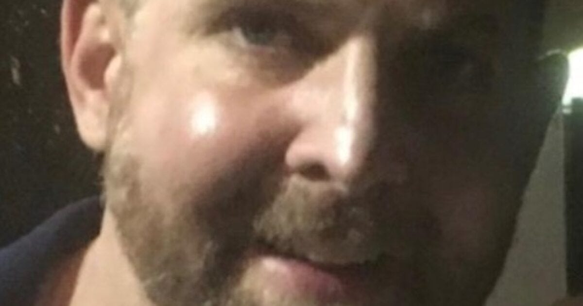 Close-up of a smiling man with a beard, captured in low light, showcasing facial features and a friendly expression.