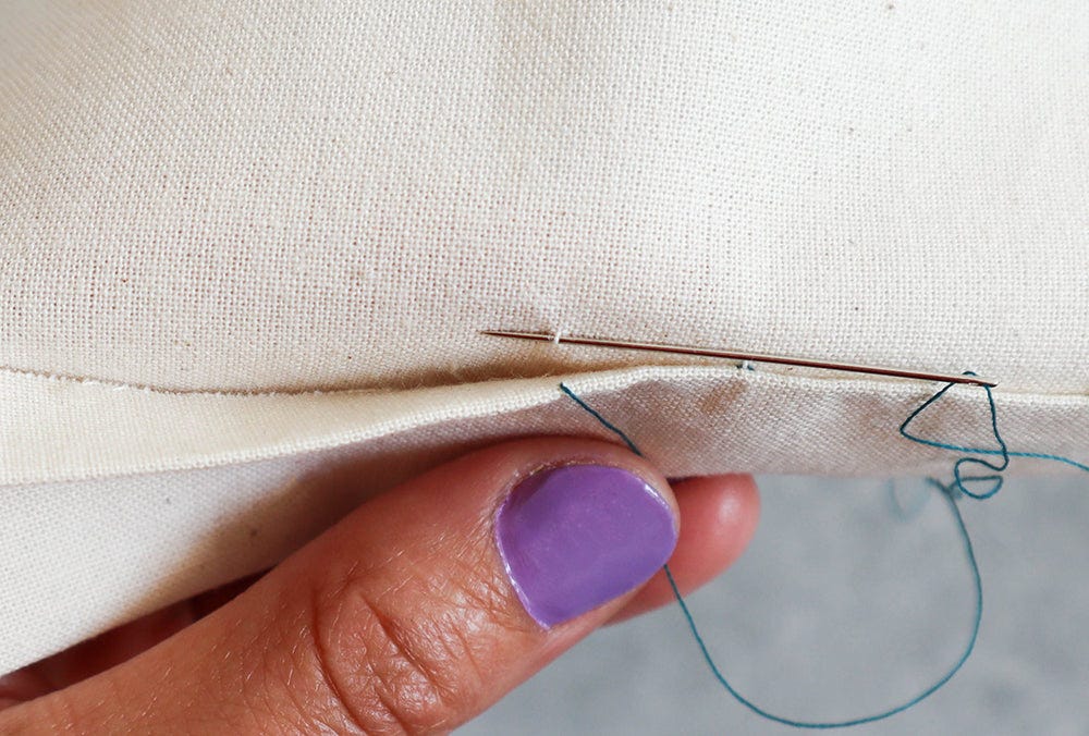 Cream muslin fabric with hem folded up, needle with teal thread sewing a slip stitch