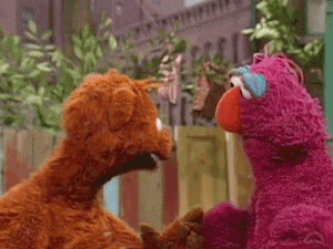 Due pupazzi pelosi di Sesame Street sono in piedi l'uno di fronte all'altro e si guardano. Il pupazzo a sinistra è di colore marrone aranciato, mentre quello a destra è rosa magenta brillante con un grande naso arancione e occhiaie azzurre. Sembrano essere impegnati in una conversazione, con il pupazzo rosa che tiene la mano o il braccio dell'altro. Lo sfondo è un set all'aperto di Sesame Street con recinzioni in legno e piante.