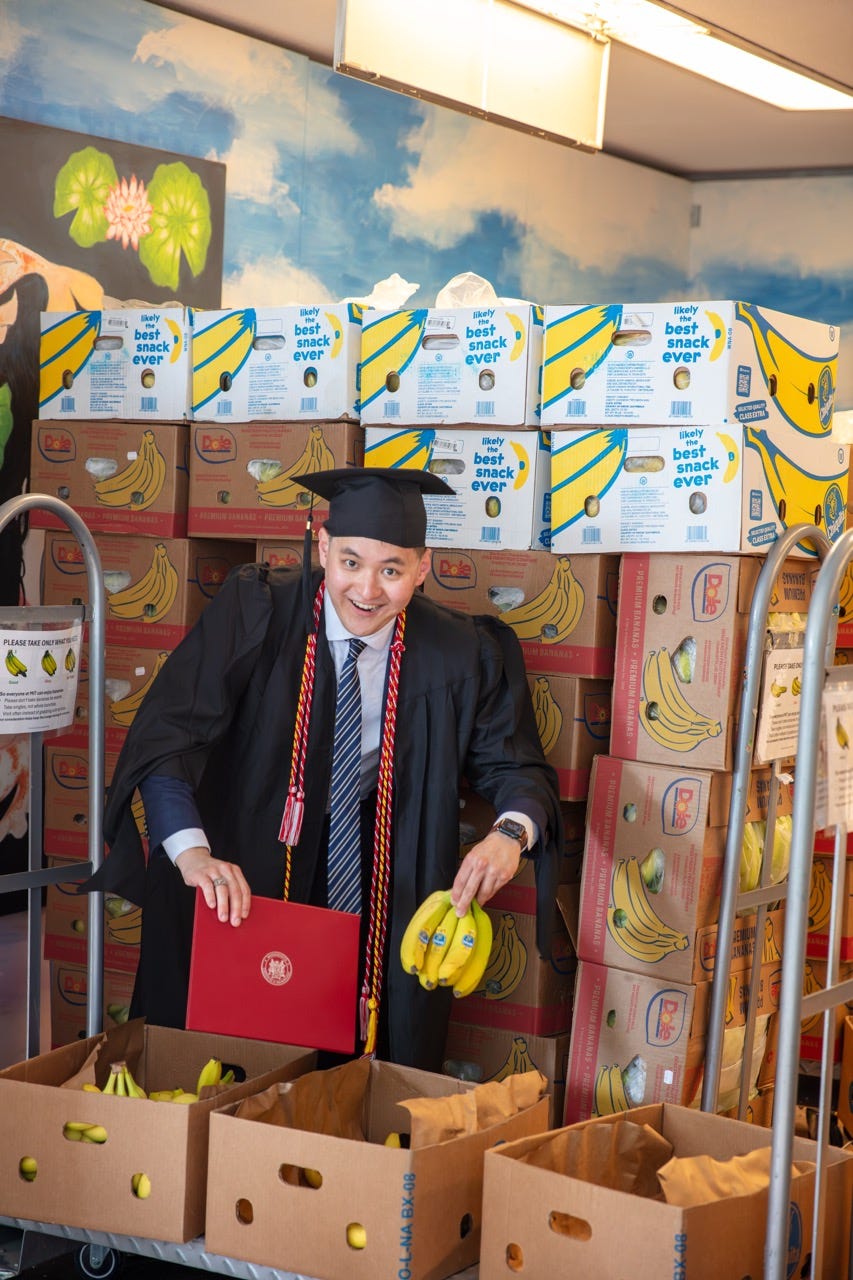 Junru in MIT graduation regalia holding bananas and a red diploma folder in front of stacked banana boxes Junru in MIT graduation regalia holding bananas and a red diploma folder in front of stacked banana boxes
